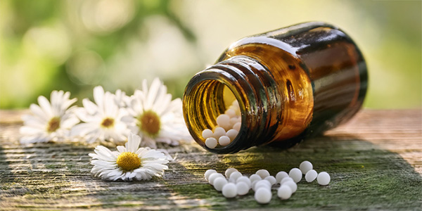 Braunes Glasfläschchen liegt auf einer Holzoberfläche, weiße Kügelchen sind herausgefallen. Daneben liegen Kamillenblüten. Darstellung weist auf homöopathische oder pflanzliche Arzneimittel hin.